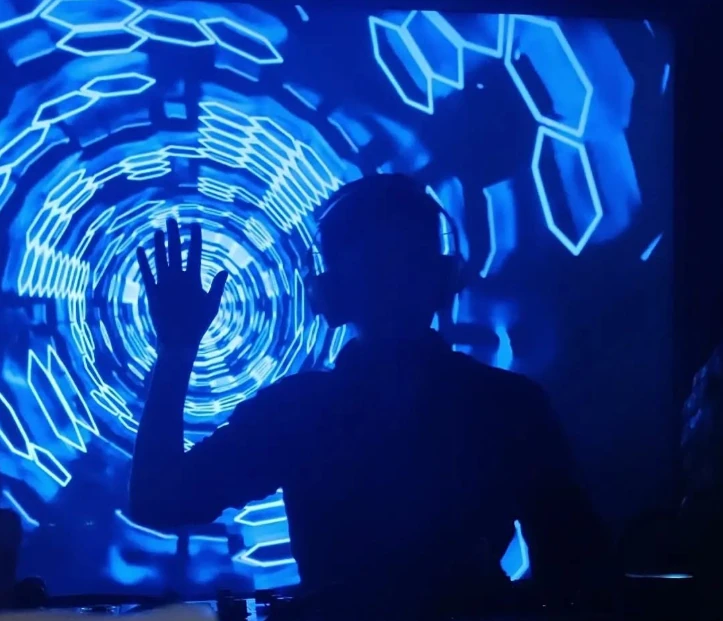 Andrés Romero tras los equipos de DJ, con proyecciones azules al fondo